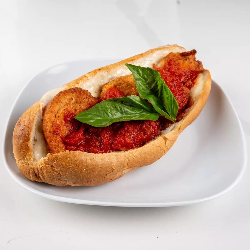 Chicken parmesan hero sandwich with melted cheese, marinara sauce, and fresh basil on a white plate.