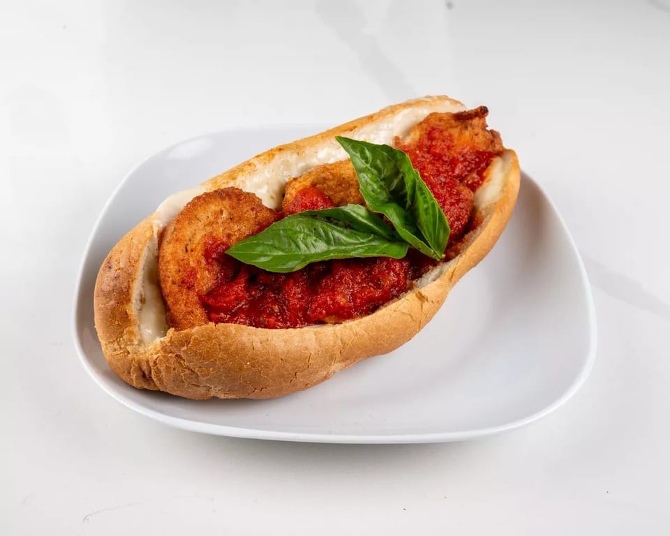 Chicken parmesan hero sandwich with melted cheese, marinara sauce, and fresh basil on a white plate.