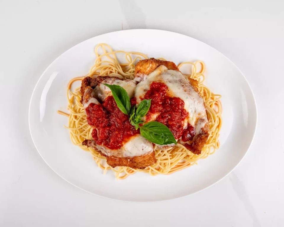 Chicken parmesan with spaghetti, marinara sauce, melted cheese, and fresh basil on a white plate.