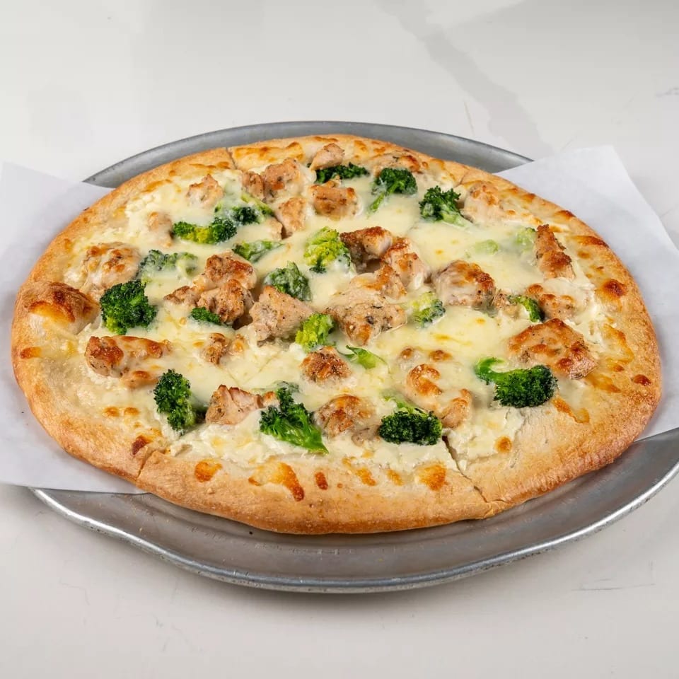 Chicken and broccoli pizza with white sauce on a metal tray, on a white marble surface.
