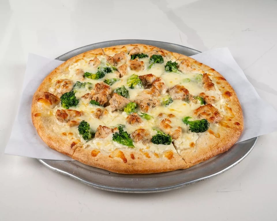 Chicken and broccoli pizza with white sauce on a metal tray, on a white marble surface.